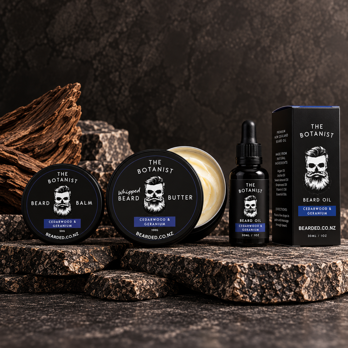 BEARDED. men's grooming set including a jar of whipped beard butter, a round tin of beard balm, a bottle of beard oil, and its retail box, arranged on dark textured stone with driftwood in the background.