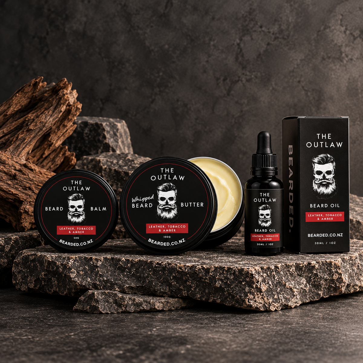 A set of beard grooming products from BEARDED. Arranged on a dark stone surface. The set includes The Outlaw Bearded Balm, a tin of Bearded Butter (open to show the cream), a bottle of Beard Oil, and its retail box.