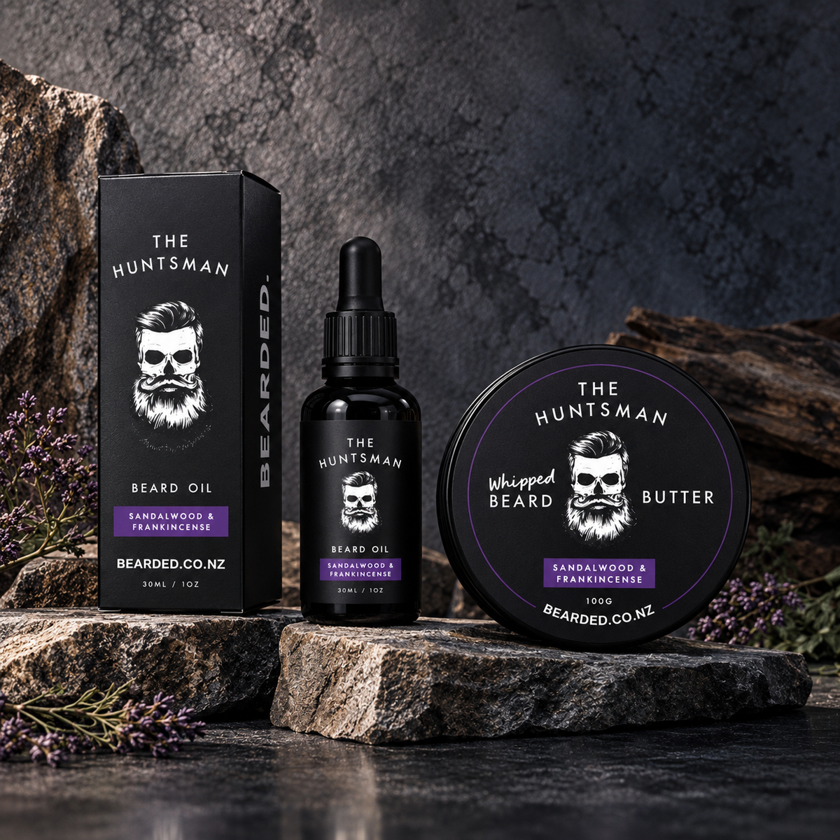 Beard grooming product set including a beard oil bottle, matching box, and round butter tin, all branded The Huntsman and Bearded Co. NZ, arranged on rocks with dried flowers against a dark textured backdrop.