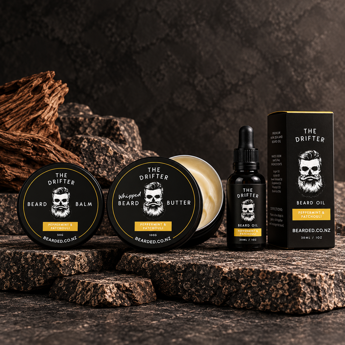 Beard grooming product set from The Drifter, including beard balm tins, whipped beard butter, a beard oil dropper bottle, and a beard oil box, arranged on dark textured stones with wood props.