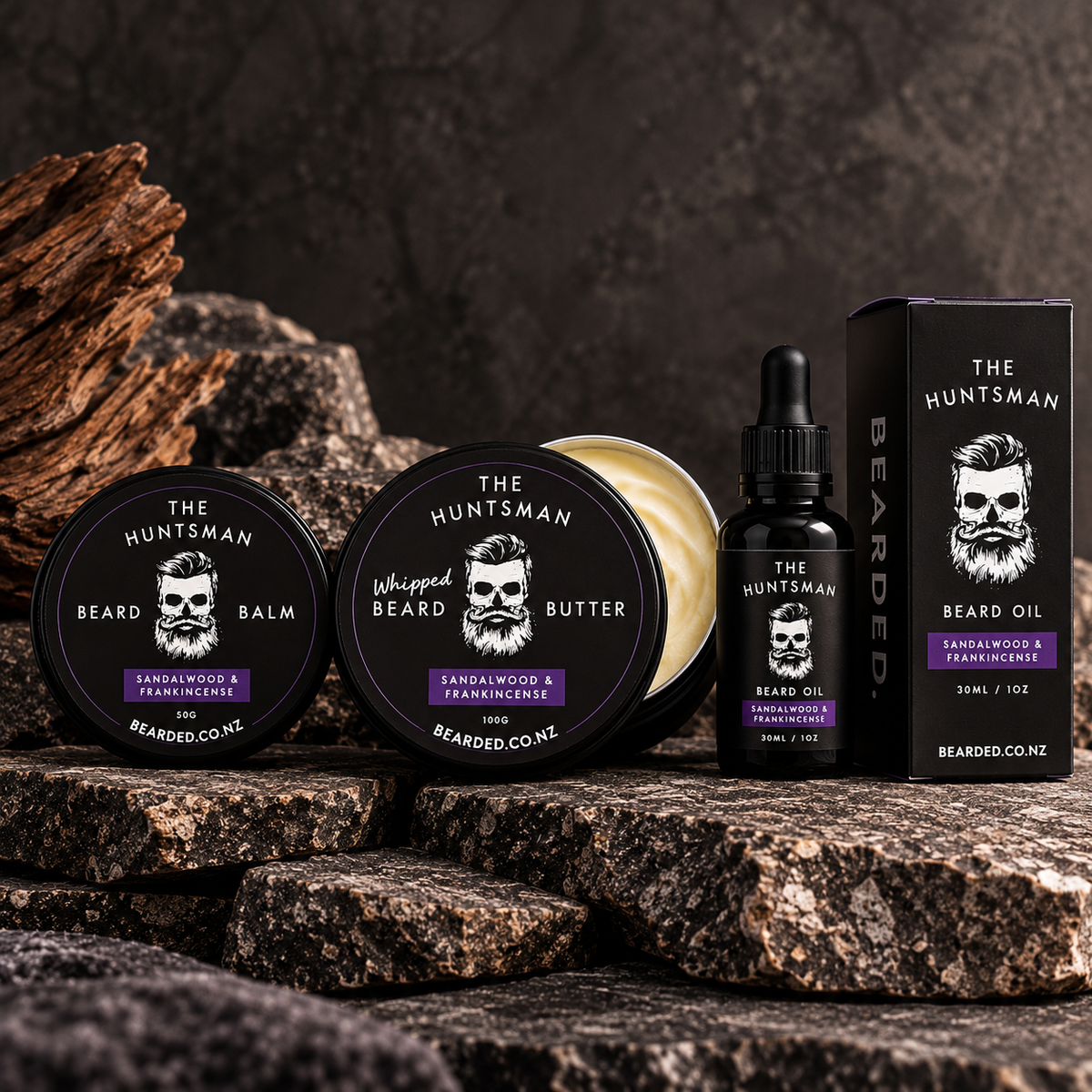 Beard grooming product set The Huntsman from BEARDED, including beard balm, whipped beard butter, beard oil, and packaging, arranged on dark rocks with wood in the background.