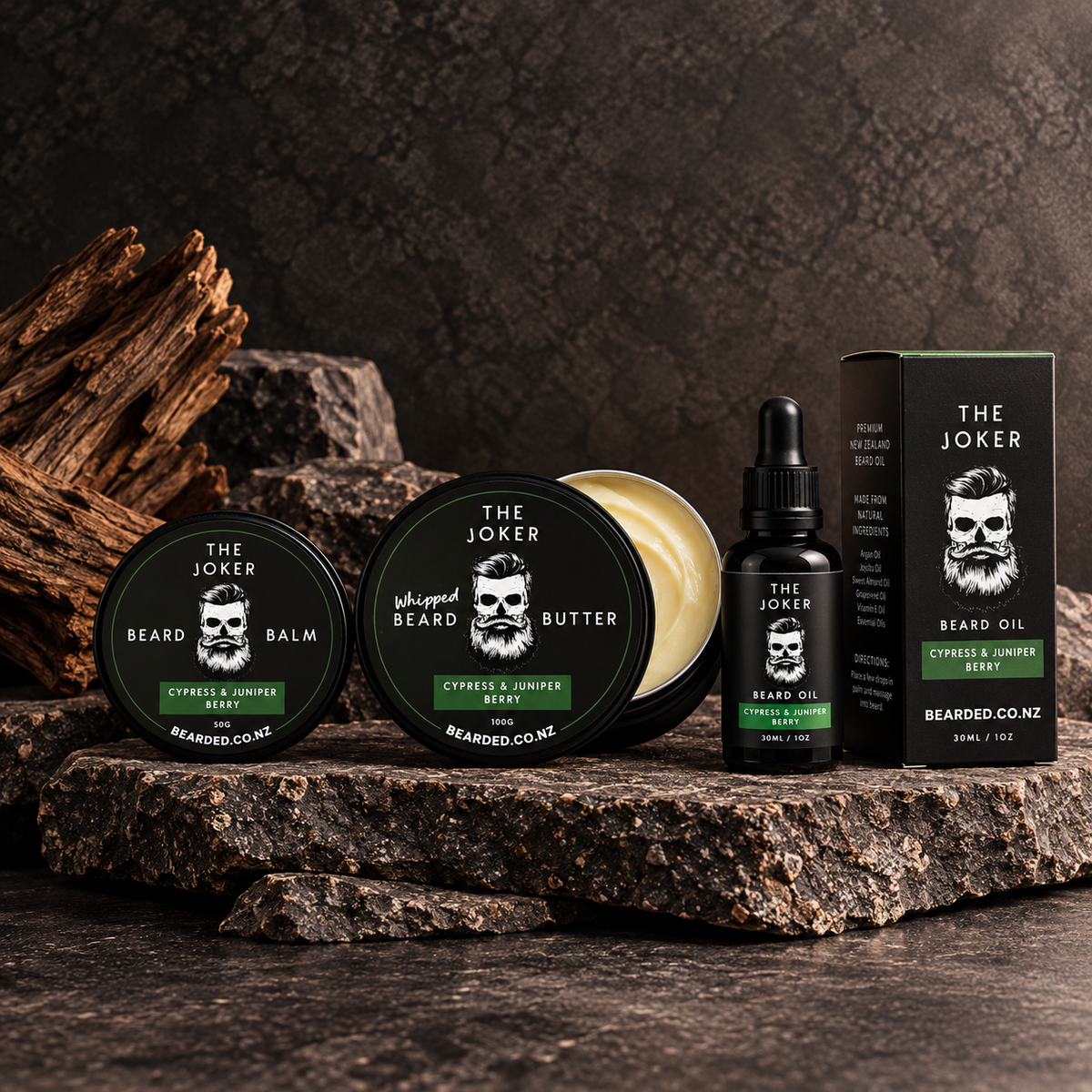 Three beard care products from The Joker are arranged on a dark rustic stone surface: a round beard balm tin, a round whipped beard butter tin with its lid off showing pale cream product inside, a small dropper bottle of beard oil, and a tall product box. The labels are black with white skull graphics and green accents, and the scent is described as Citrus & Juniper Beard.