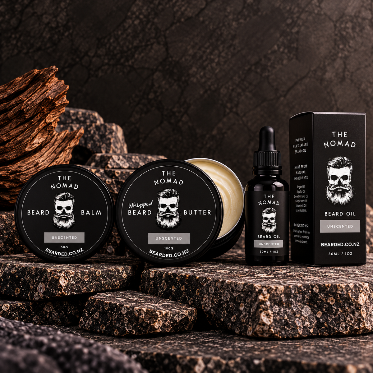 A beard grooming set from The Nomad, including an uncentred beard balm tin, whipped beard butter jar, a bottle of beard oil, and a beard oil box, arranged on dark textured stone blocks.