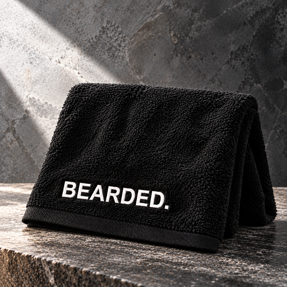 A black textured towel with 'BEARDED.' embroidered in white capital letters, folded and displayed on a dark stone surface against a gray stone wall.